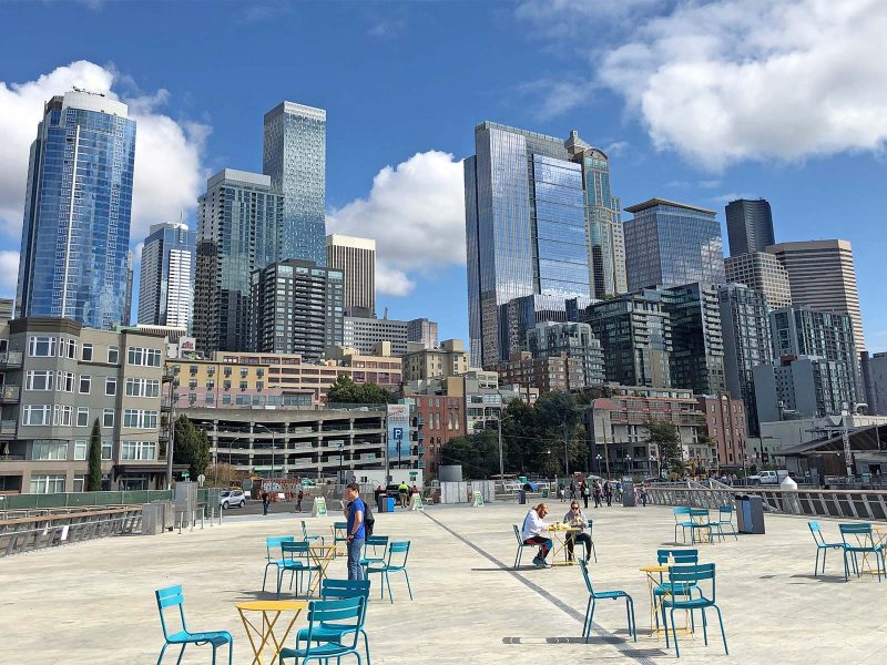 Pier 62 in downtown Seattle: People sitting at bistro tables and chairs on a sunny day, cityscape in background. Blue sky with clouds.
