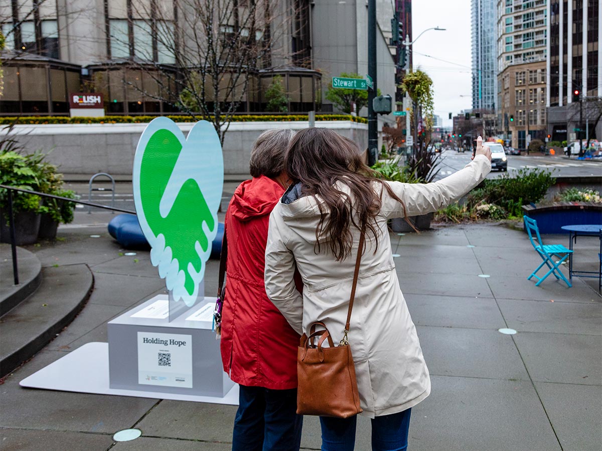 Two people taking a selfie in front of metal sculpture in McGraw Square: Light blue and green hands in the shape of a heart