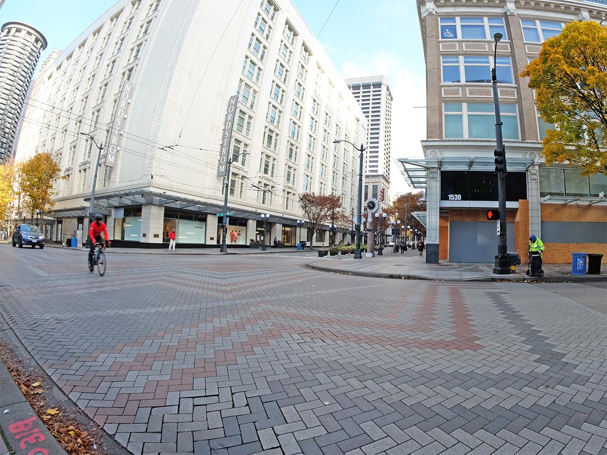 A quiet, mostly empty street in downtown Seattle: one biker, a MID ambassador, one car. Brick road with greys and reds. Tall white building and blue sky.