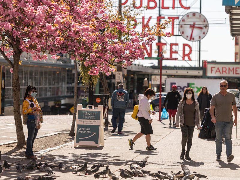 Visitors to Pike Place Market walking around on a sunny day. Cherry blossoms, pigeons, Pike Place Market Center sign.