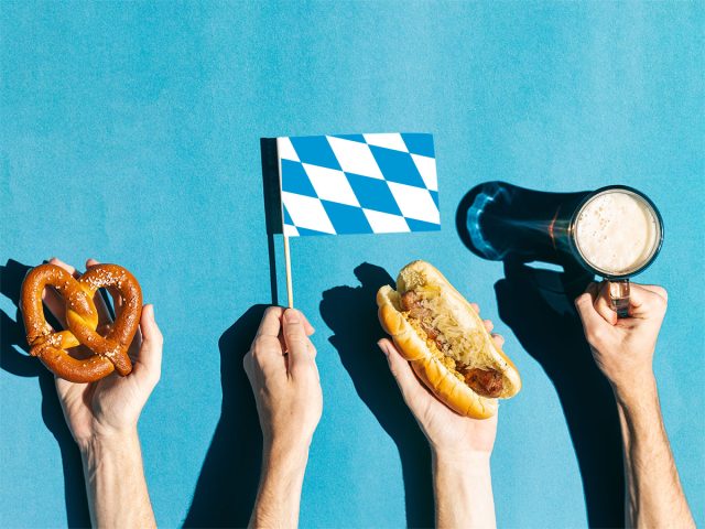 Hands holding Oktoberfest objects: pretzel, Bavarian flag, hot dog and beer against a blue background