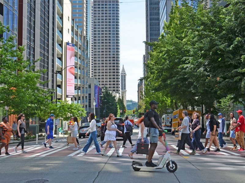 Pedestrians and scooter on a sunny summer day in a downtown Seattle crosswalk. Cityscape in the background.