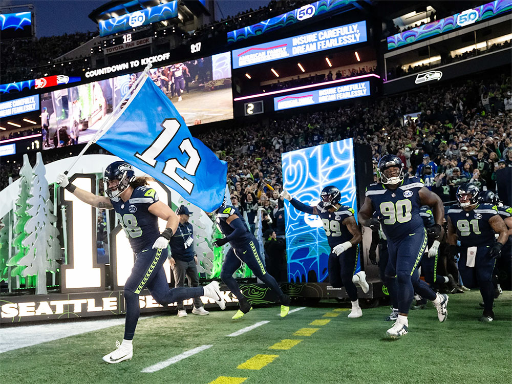 Seattle Seahawks running out onto Lumen Field. The player in front is waving a "12th Man" flag.